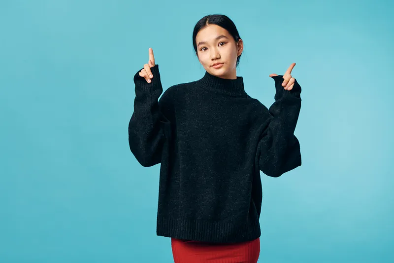 Young woman wearing black sweater and red skirt standing with fingers pointing up on blue background. Casual outfit, confident pose and studio portrait with copy space on side.