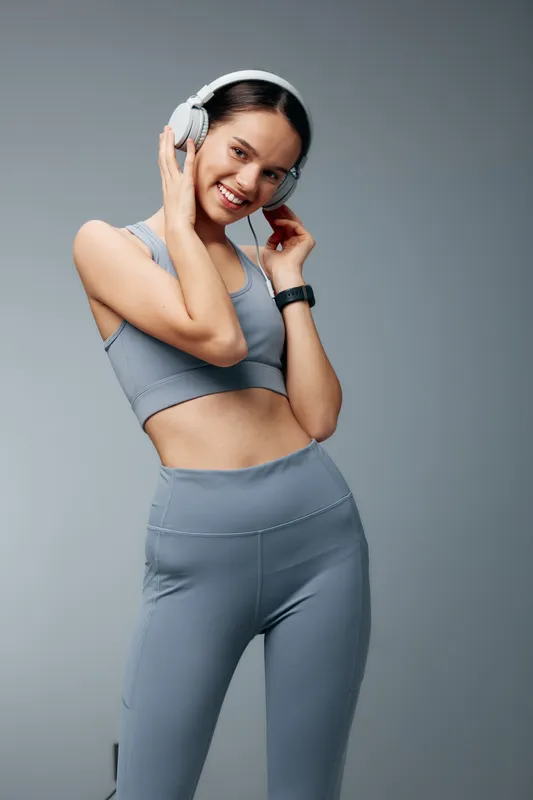 Fitness woman wearing a gray sports set with headphones, smiling in a clean studio, showcasing an active lifestyle, athletic fashion and energy during a workout moment.