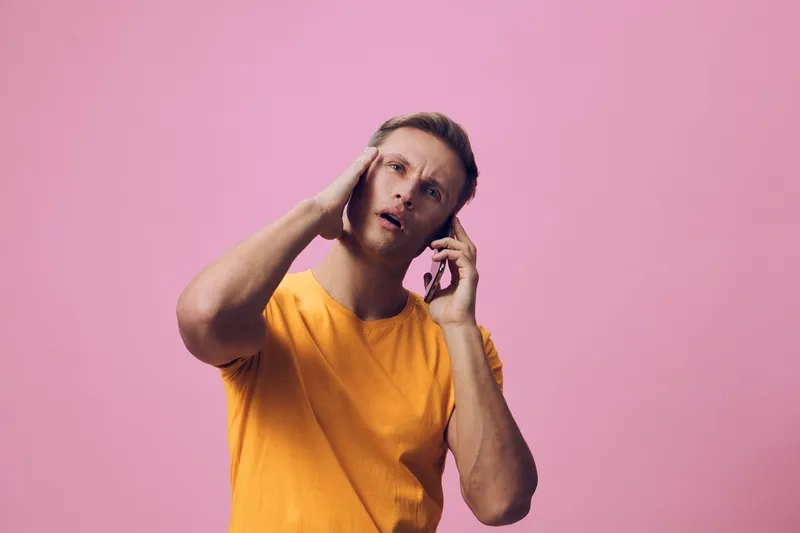 Confused man wearing yellow t-shirt, holding phone to ear, looking puzzled with furrowed brows, isolated on pink background. People emotional reaction concept