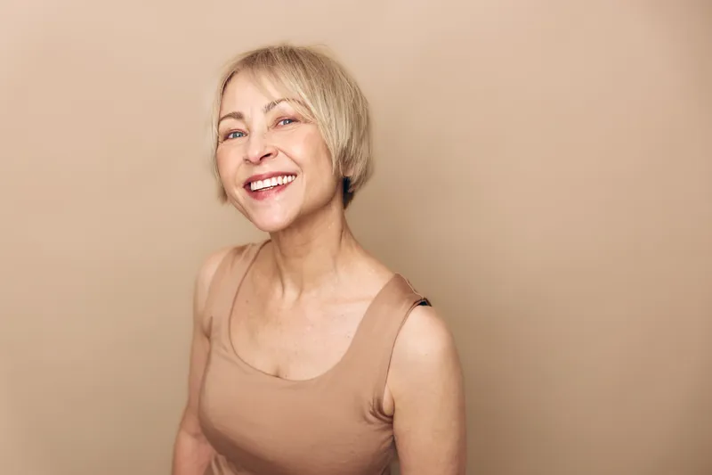 Smiling mature woman with short blond hair wearing beige sleeveless top looks happy and confident against plain beige background. Concept of beauty, aging, positivity, and wellness.