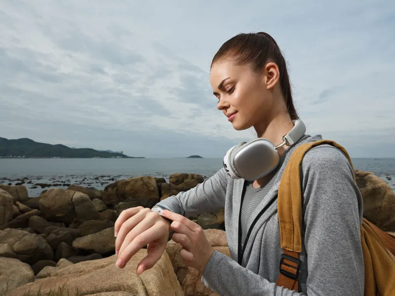 Young woman with headphones checking smartwatch by the seaside, showcasing a sporty style and a relaxed atmosphere with natural tones