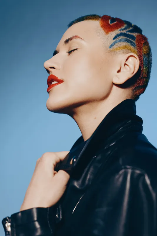 Woman with colorful shaved hair and red lipstick posing confidently in black leather jacket against a blue background, showcasing edgy alternative style.