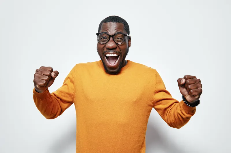 African man with dark skin wearing glasses and orange sweater celebrating success with raised fists and joyful expression on white background. Excited, happy and energetic.