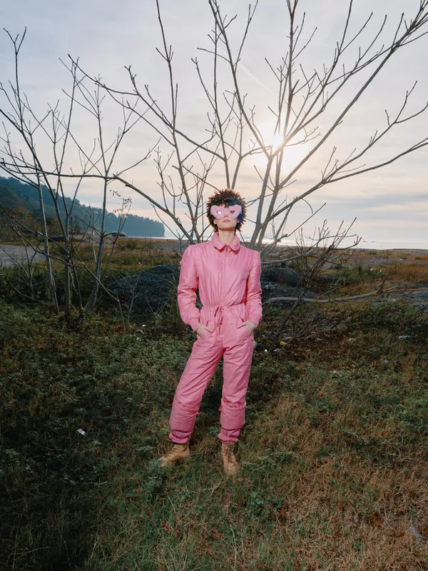 Person outdoors wearing a pink jumpsuit and mask stands beside a bare tree in a wide countryside landscape, low sun on the horizon, creating a mysterious and atmospheric scene.