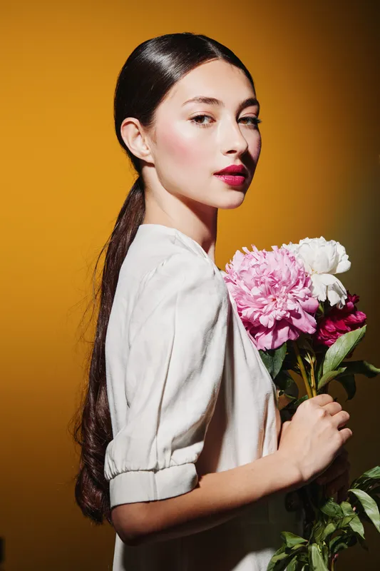 Elegant young woman with sleek black hair poses in white blouse holding bouquet of flowers against bright yellow background. Concept for beauty creative fashion portrait.