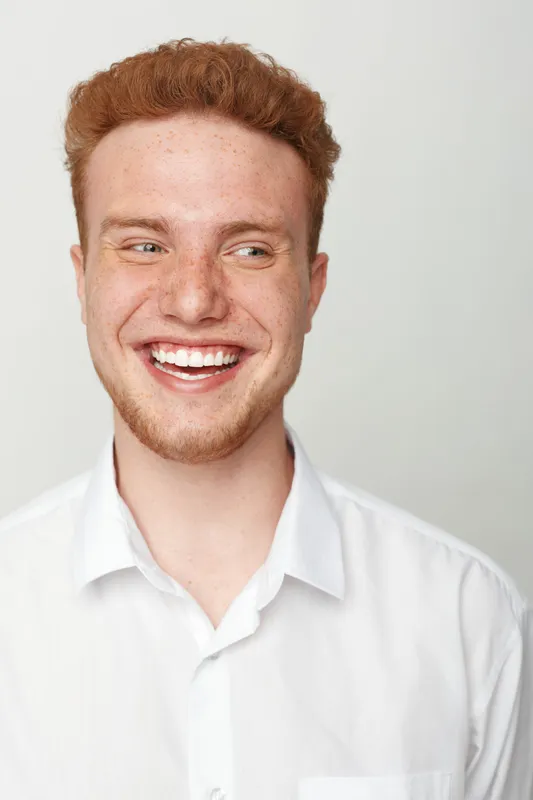 Smiling young man with red hair and beard wearing white shirt looks away with happy expression. Studio portrait on light background. Concept of joy, youth, and positive emotions.
