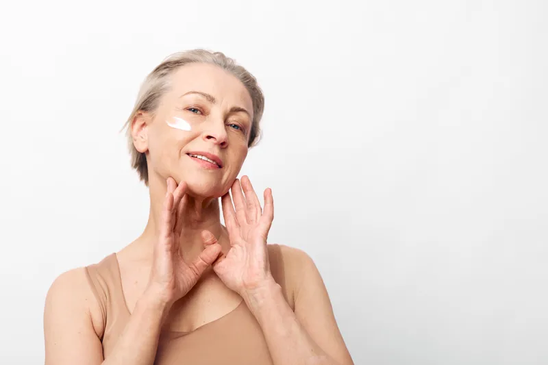 Mature woman with light hair applies cream under eyes, smiling gently. Skincare routine concept for aging skin, beauty care, and self-confidence in natural light.