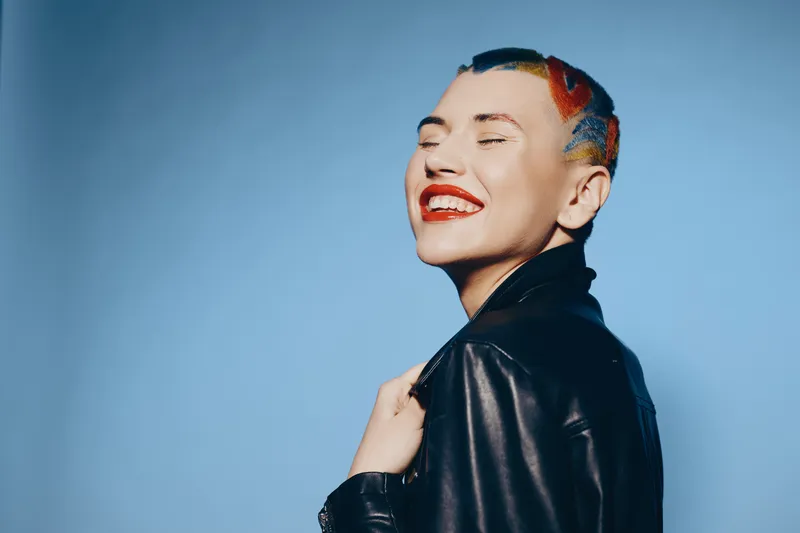 Smiling woman with colorful short hair wearing a black leather jacket, expressing happiness and confidence against a simple blue background in studio lighting.
