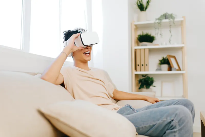 Молодой человек погружен в опыт виртуальной реальности: он улыбается в VR-очках, удобно сидя на светлом диване в светлой уютной гостиной, наполненной растениями и книгами