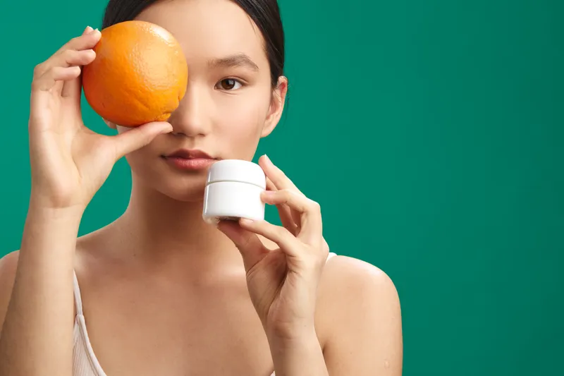Asian woman holds an orange beside her face while presenting a small cosmetic jar, set against a vivid green backdrop to convey fresh beauty and playful wellness.