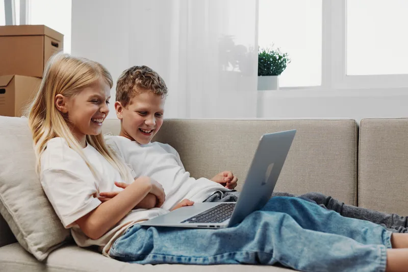 Two happy children sitting on a couch using a laptop, smiling and enjoying digital content together at home.