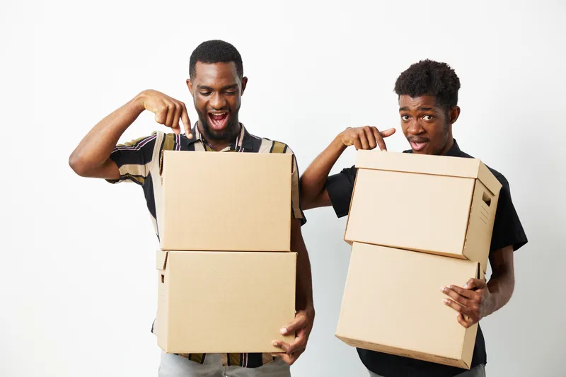 Happy black men celebrating a successful move while holding cardboard boxes, showcasing their excitement and teamwork in an indoor setting.