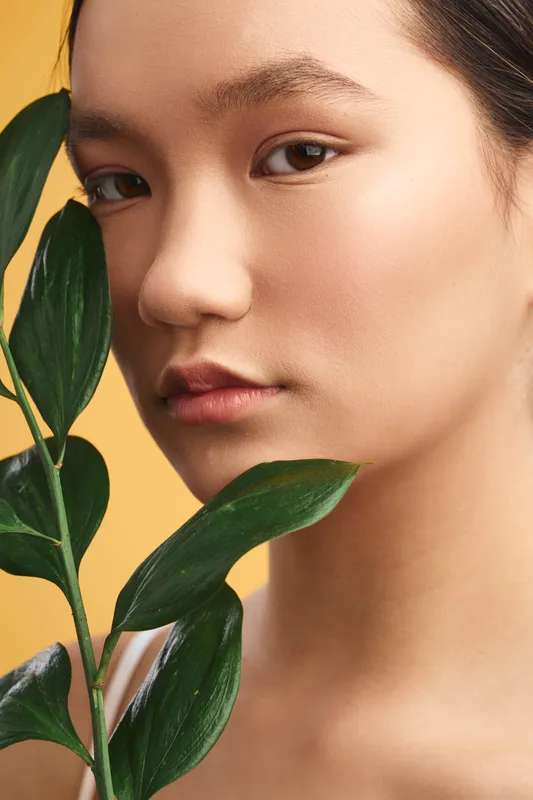 Asian woman gazes softly at the camera in a close up portrait, with leafy plants framing her face and a warm backdrop, conveying calm, natural beauty, and quiet elegance.
