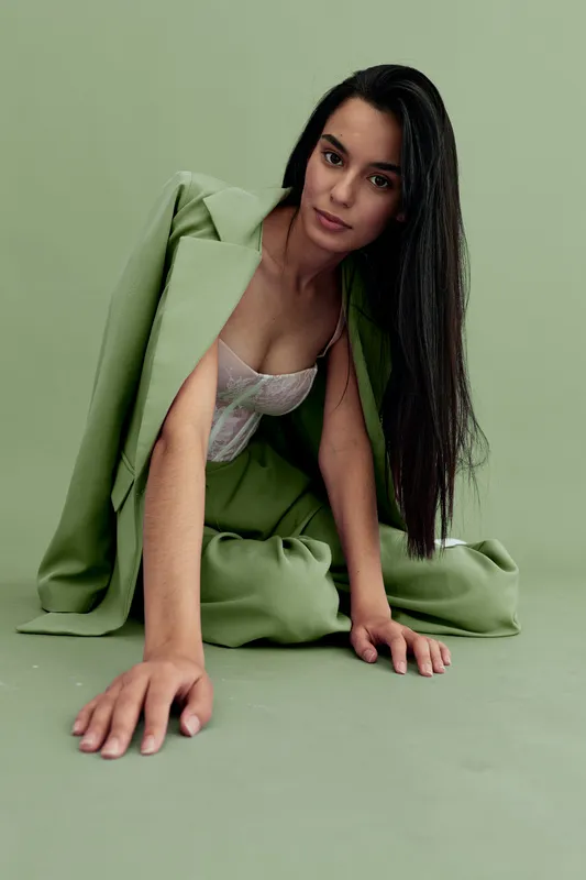 Young woman wearing a pastel green suit and lace top poses on a matching green floor and background, showcasing modern fashion with a calm, poised expression.