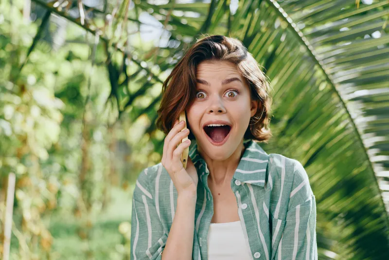 Young woman outdoors with a wide surprised expression among green tropical plants, wearing a striped shirt, capturing a vibrant, playful mood.