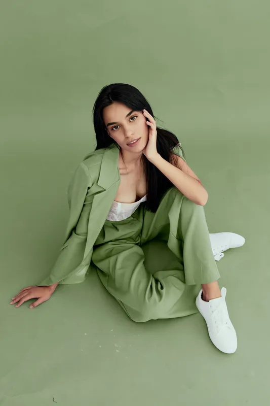 stylish woman in green suit sitting on floor against matching background, casual white sneakers, dark hair, relaxed pose, contemporary fashion concept