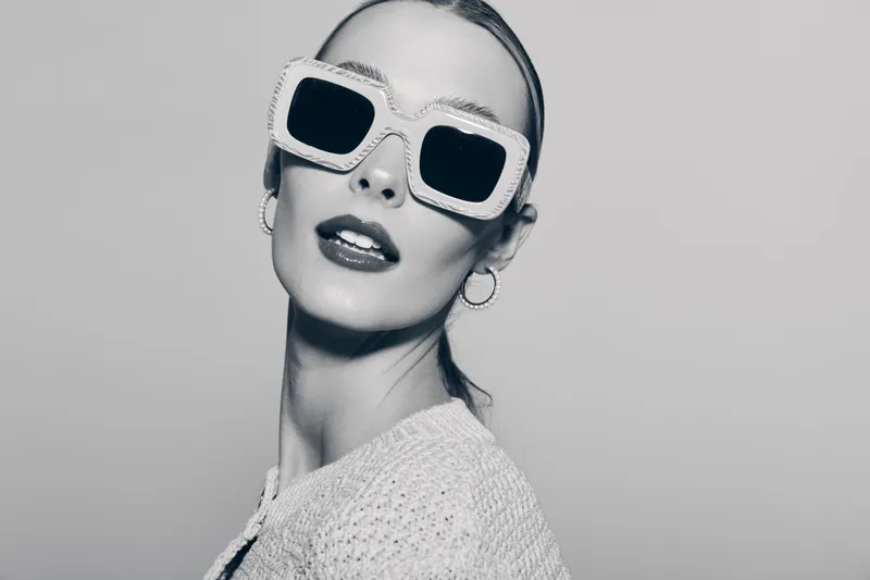 Portrait of a fair skinned white woman wearing stylish large sunglasses and hoop earrings with slicked back hair in a black and white studio image.