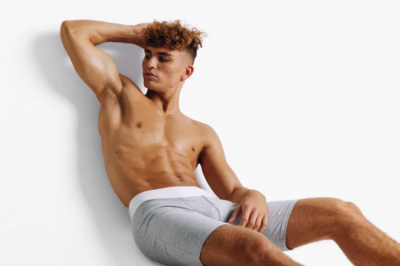 Young man with curly hair posing shirtless in gray shorts against white background, showing muscular body and relaxed confident expression in studio setting.