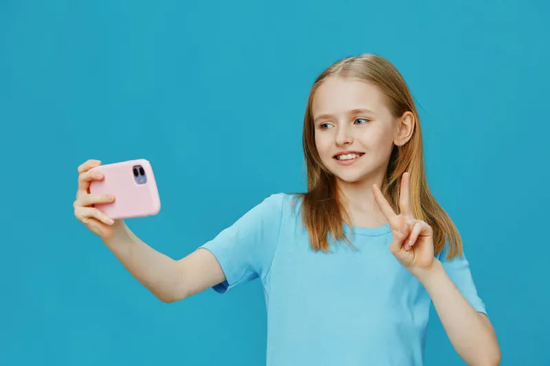 Joyful girl taking a selfie with a smartphone against a vivid blue background, wearing a casual blue t shirt and showing a peace sign, expressing happiness and confidence