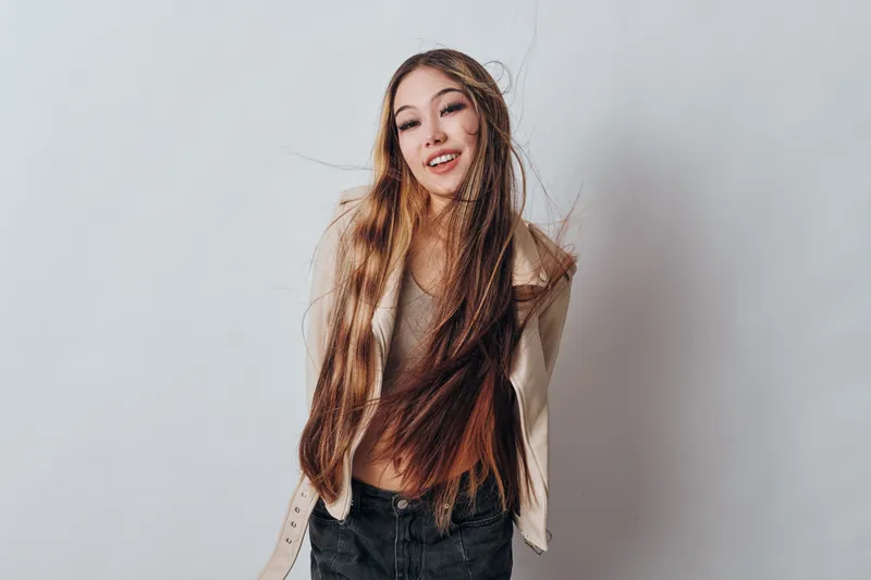 Young woman with long straight hair smiling standing against light background wearing casual beige jacket and dark jeans in studio portrait beauty fashion concept.