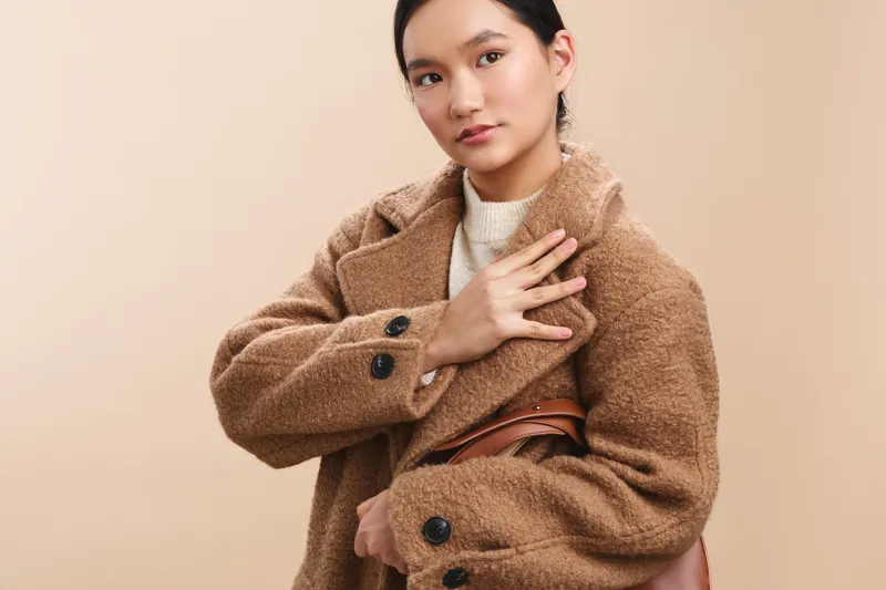 Asian woman wearing a tan cozy coat stands confidently in a warm studio setting, conveying calm professionalism and contemporary style.