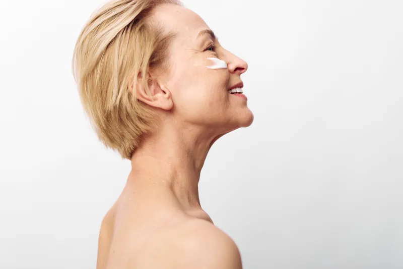 Smiling middle aged woman with short blonde hair applies facial cream on cheek, isolated on white background. Concept of skincare, beauty routine, anti aging and self care.