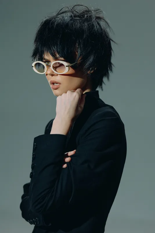 Stylish woman with short black hair and white sunglasses wearing a black outfit in a minimalistic setting.