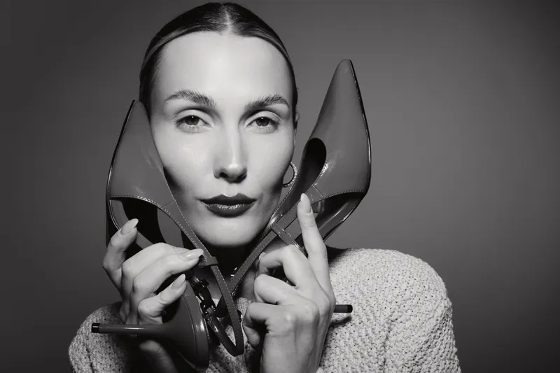 Caucasian woman with pale skin holds red high heels near face looking confidently at camera. Black and white portrait with focus on fashion accessories and female beauty concept.