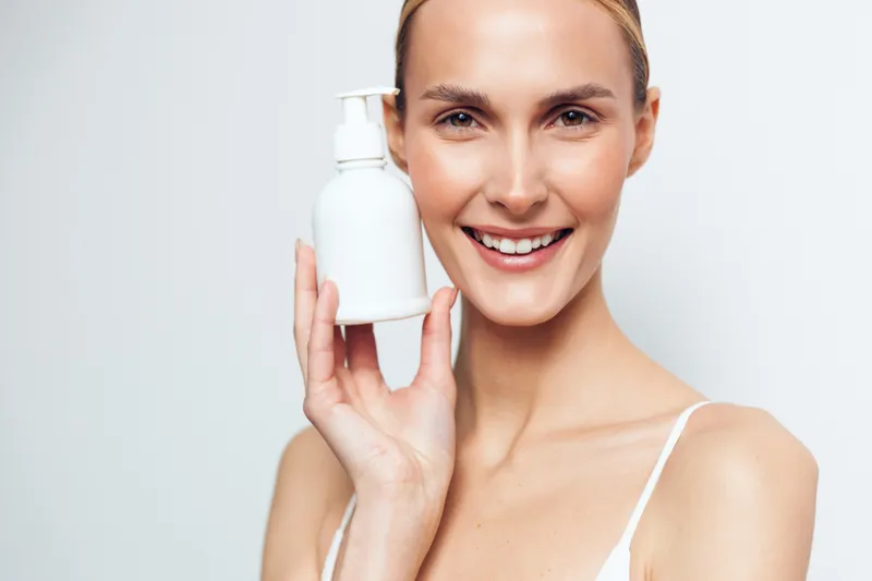 Smiling woman with clear skin holding white cosmetic bottle near face. Beauty and skincare concept with natural makeup, soft lighting, and light background. Healthy lifestyle and wellness.