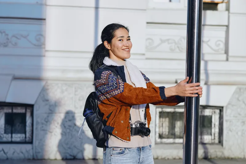 Smiling woman outdoors with a backpack by a metal pole next to a light building, wearing a patterned jacket and scarf, urban daytime style, casual street fashion, travel mood and friendly energy