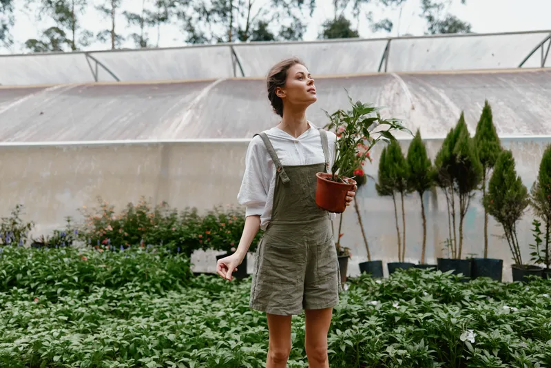 Young woman in casual overalls holds a potted plant in a lush greenhouse, expressing joy amidst vibrant greenery