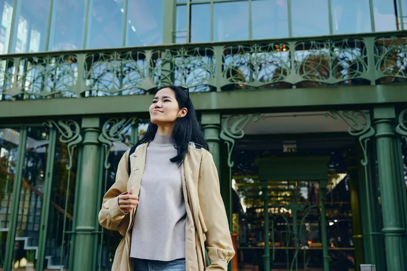 Woman standing outdoors near a glass building, wearing a beige coat and gray sweater, looking upward with a calm expression, urban fashion shot highlighting street style and modern architecture