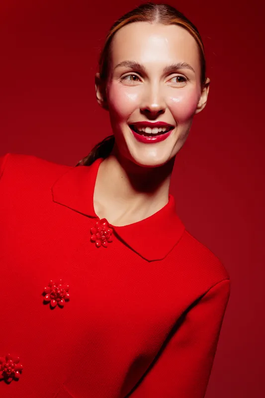 Smiling woman in red sweater with decorative buttons poses against red background. Bright makeup and natural light highlight her joyful expression and smooth skin tone.