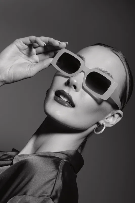 Stylish white woman with light skin wearing large sunglasses and hoop earrings poses with hand near face in black and white studio portrait. Concept of fashion, elegance, and accessories.