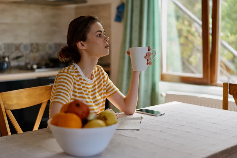 Woman relaxing at home, sits at a wooden kitchen table, holds a mug, soft morning light from a window, bowl of fruit nearby, casual lifestyle scene with serene mood