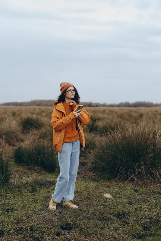 Woman outdoors walking along a grass field path, wearing orange jacket and jeans, beanie hat for cool autumn, calm lifestyle moment, cloudy sky, natural light, relaxed gaze