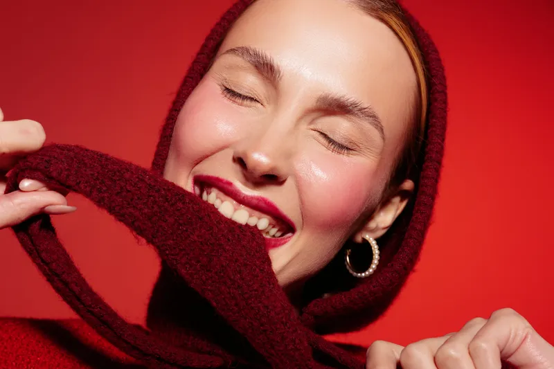 Smiling woman with closed eyes wearing a red hooded sweater on red background. Close-up portrait with natural makeup and joyful expression. Concept of happiness and cozy fashion.