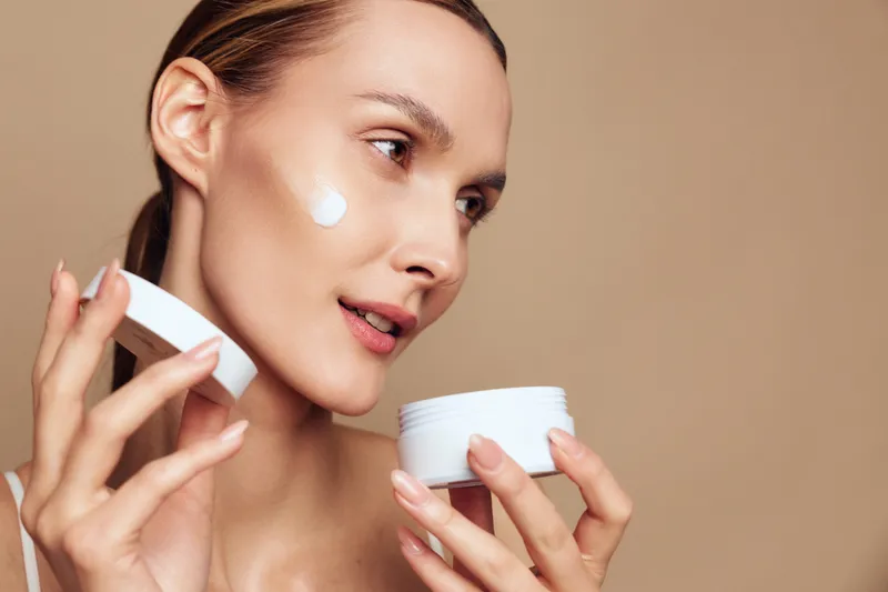 Young woman applies face cream on cheek holds open jar of moisturizing skincare product looks focused and relaxed isolated on beige background with natural makeup and smooth skin