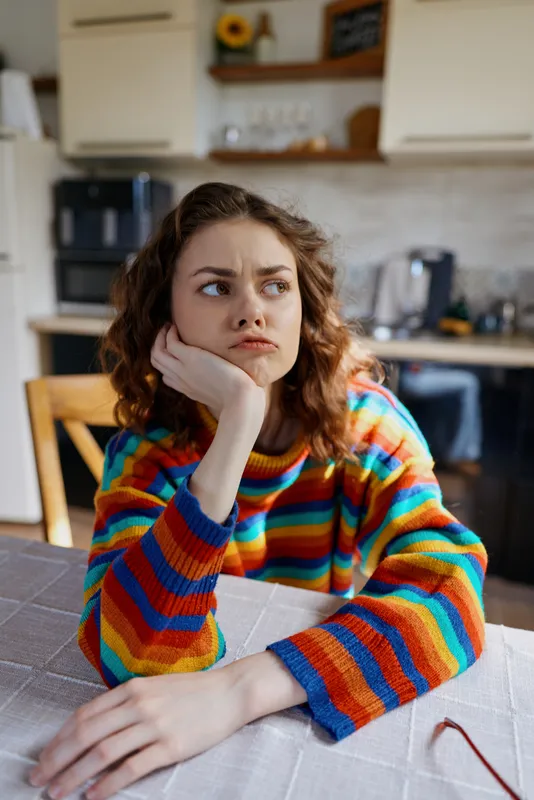 A woman in a rainbow colorful sweater sits at a kitchen table, chin resting on her hand, looking pensive. Bright morning light in a cozy home scene conveys everyday mood and contemplation.
