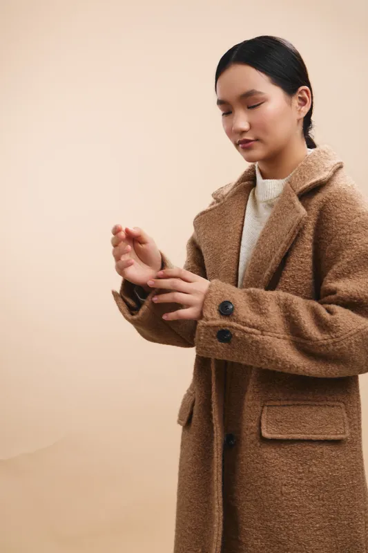 asian woman wears a warm brown coat and adjusts her sleeve in a neutral studio, conveying calm focus and professional poise with understated style and a thoughtful presence.