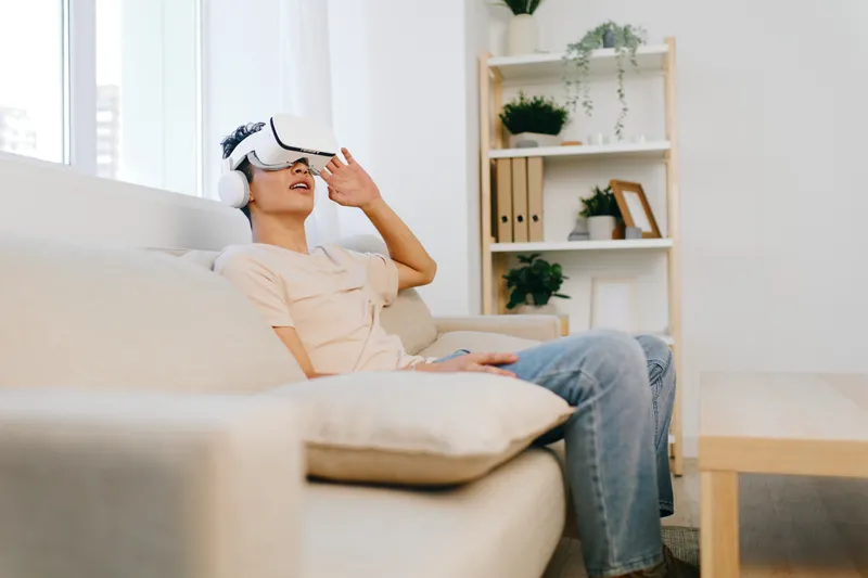 Молодой человек погружен в опыт виртуальной реальности дома: он в шлеме VR, удобно сидит на мягком диване в светлой современной гостиной со стильным декором и зелеными растениями