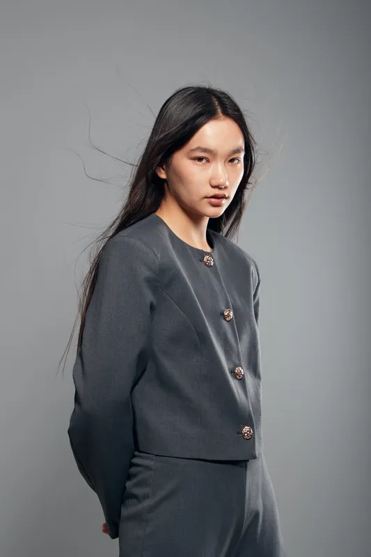 Woman with long hair wearing dark grey suit posing in studio with neutral gray background and soft lighting. Professional portrait with confident expression and minimalistic style.