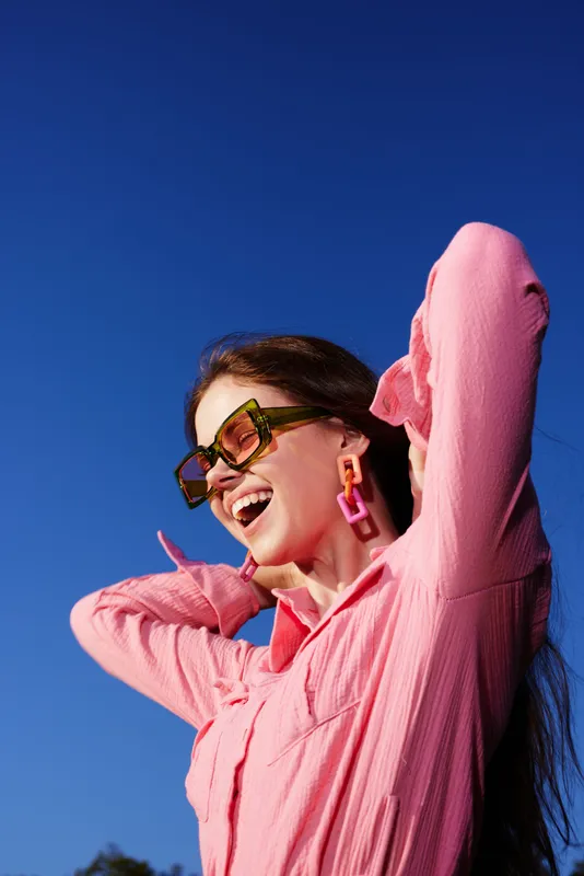 Joyful young woman in pink shirt and sunglasses enjoying sunny day against blue sky, capturing happiness and carefree lifestyle in vibrant summer fashion
