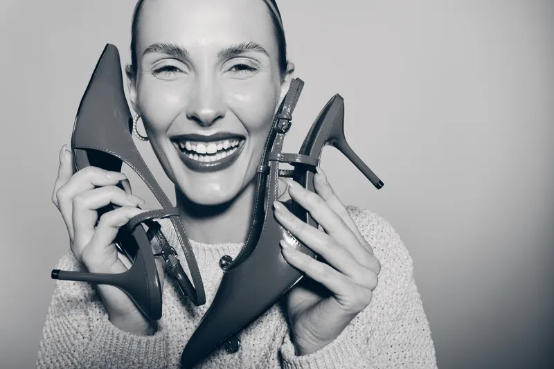 Happy Caucasian woman with light skin holding high heel shoes smiling in studio. Concept of fashion, footwear, joy and lifestyle.