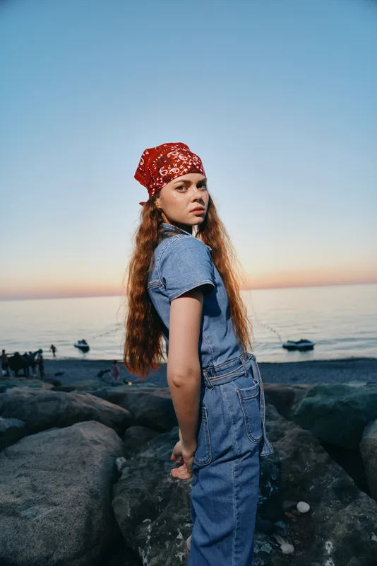 lifestyle woman in modern boho-western denim street style with long red hair and red bandana standing by rocks at sunset, film color warm tones