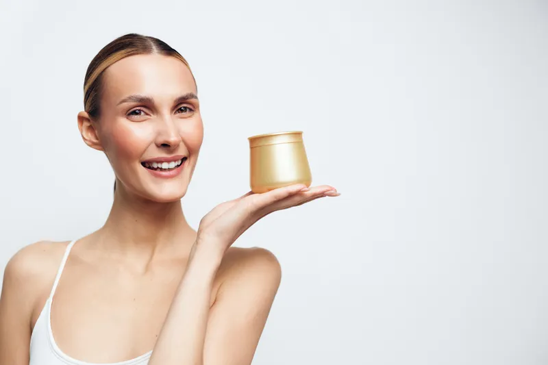 A smiling woman with clear skin holds a golden cosmetic jar on her hand against a white background. Concept for skincare, beauty, wellness, and natural cosmetics.
