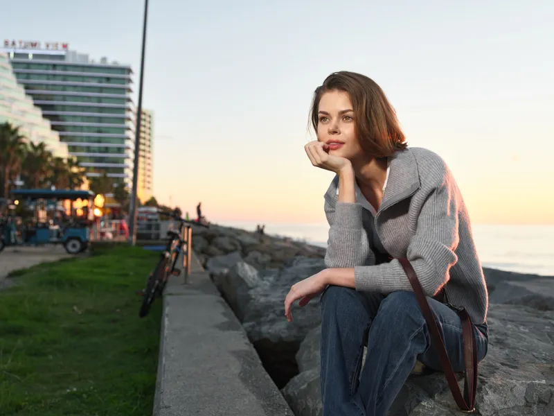 A thoughtful woman sits on a concrete seawall at sunset, resting her chin on her hand, gazing toward the horizon with a calm and contemplative mood.