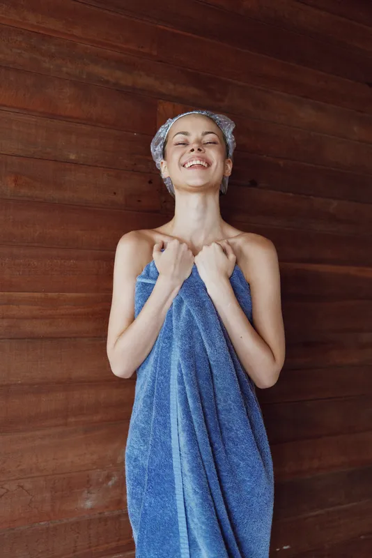 smiling woman wrapped in blue towel with headscarf stands against wooden wall, expressing happiness and relaxation after bath or spa treatment in warm natural light