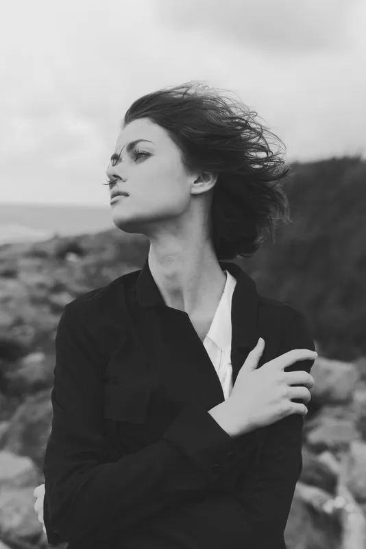 Moody black and white image of a windswept woman standing on a rocky coastline, wearing a dark jacket, with a thoughtful gaze beneath a dramatic sky.