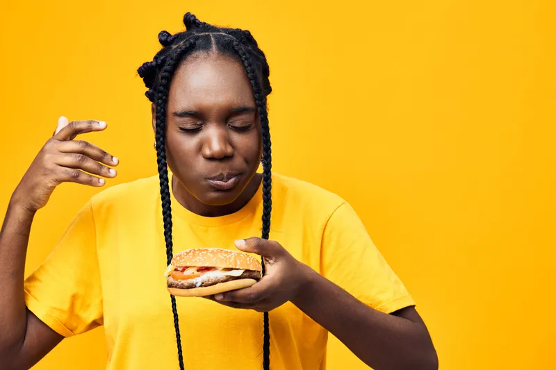 Young woman enjoying a delicious burger against a vibrant yellow background, wearing a bright yellow t shirt, showcasing her love for tasty food in a joyful moment